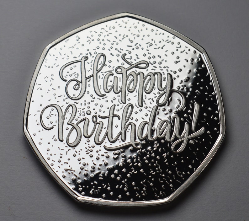 以下が含まれることがあります： 「Happy Birthday!」と書かれた銀色のコイン。コインは独特の多角形の形状をしており、小さな散らばったディテールを持つテクスチャード加工の背景があります。デザインは鮮明で詳細です。