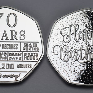 May include: A silver coin with a heptagonal shape. One side features the text "70 YEARS" and a list of time units, including "7 DECADES", "840 MONTHS", "3653 WEEKS", "25,568 DAYS", "613,920 HOURS", and "36,817,200 MINUTES". The other side features the text "Happy Birthday!" in a decorative font.