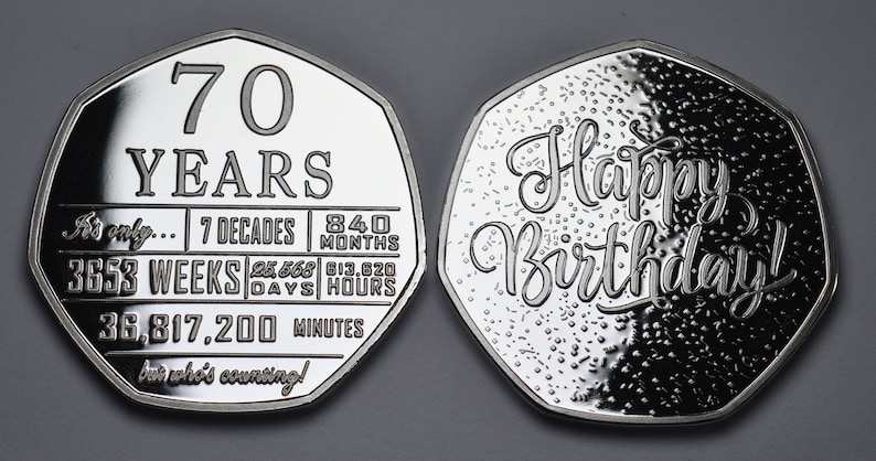 May include: A silver coin with a heptagonal shape. One side features the text "70 YEARS" and a list of time units, including "7 DECADES", "840 MONTHS", "3653 WEEKS", "25,568 DAYS", "613,920 HOURS", and "36,817,200 MINUTES". The other side features the text "Happy Birthday!" in a decorative font.