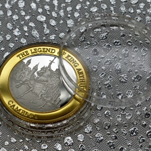 May include: A silver coin with a gold rim featuring a depiction of King Arthur on horseback. The coin is in a clear plastic case. The coin reads "The Legend of King Arthur Camelot".