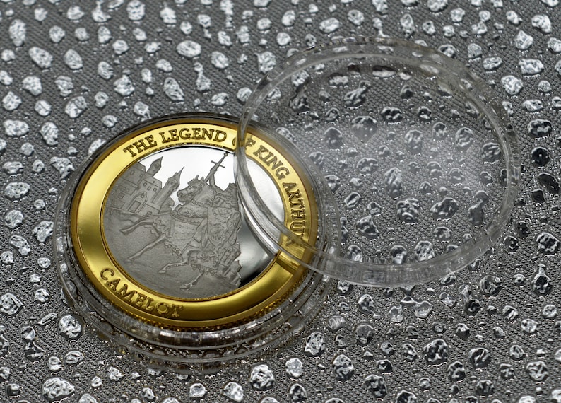 May include: A silver coin with a gold rim featuring a depiction of King Arthur on horseback. The coin is in a clear plastic case. The coin reads "The Legend of King Arthur Camelot".