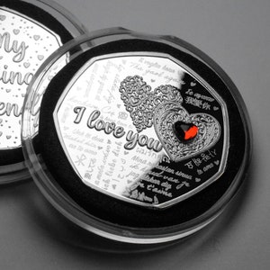 Puede incluir: Una moneda de plata con un diseño de corazón y las palabras "Te amo" en varios idiomas. La moneda está en una caja de plástico transparente con un forro de terciopelo negro.
