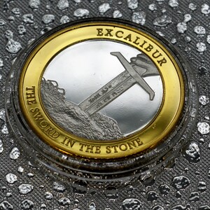 May include: A silver coin with a gold rim featuring a sword in a stone with the text "Excalibur The Sword In The Stone" around the edge.