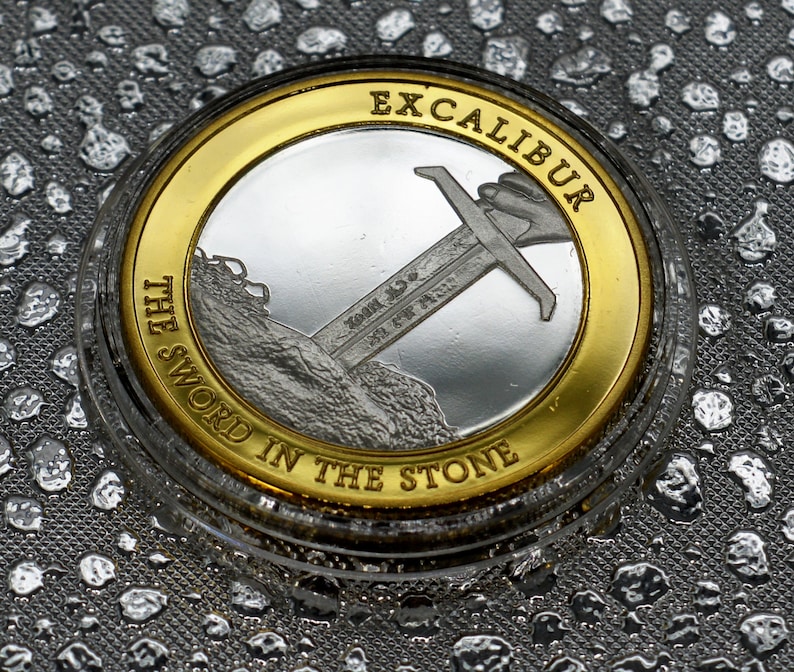 May include: A silver coin with a gold rim featuring a sword in a stone with the text "Excalibur The Sword In The Stone" around the edge.