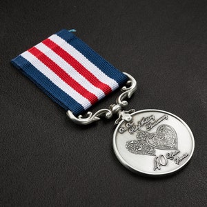Może przedstawiać: Srebrny medal z czerwono-białym wzorem serca i napisem "On Our Ruby Wedding Anniversary 40 Years Service". Medal jest przymocowany do niebiesko-biało-czerwonej wstążki.
