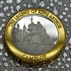 May include: A gold and silver coin with the inscription "The Legend of King Arthur Camelot". The coin features a depiction of King Arthur on horseback, riding towards a castle.