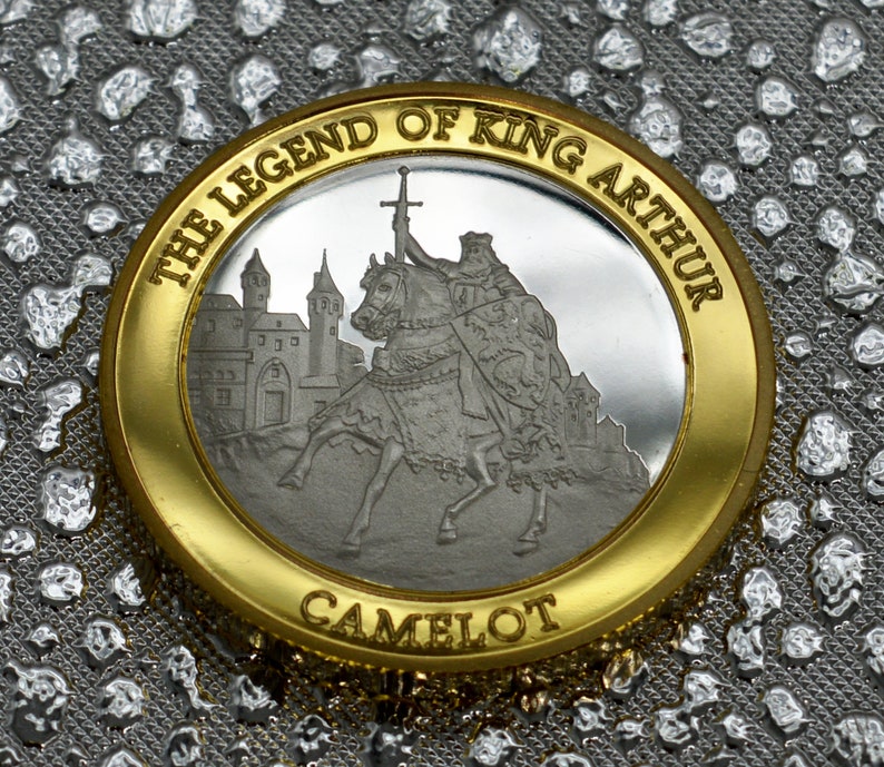 May include: A gold and silver coin with the inscription "The Legend of King Arthur Camelot". The coin features a depiction of King Arthur on horseback, riding towards a castle.
