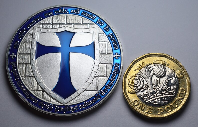 K&ouml;nnte beinhalten: Eine Silberm&uuml;nze mit einem blauen Kreuz auf einem Schild und dem Text "non adfile tuumine. Hun ix, xe nominl tuo an gloria - peugeres commilitumes cruist templique salomunici nicrust/emitonis" am Rand. Eine Goldm&uuml;nze mit einer Distel, einer Rose und einem Kleeblatt und dem Text "ONE POUND" ist ebenfalls zu sehen.
