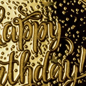 May include: A gold-coloured card with the words "Happy Birthday" embossed in a decorative script font. The background is textured with a pattern of small squares.