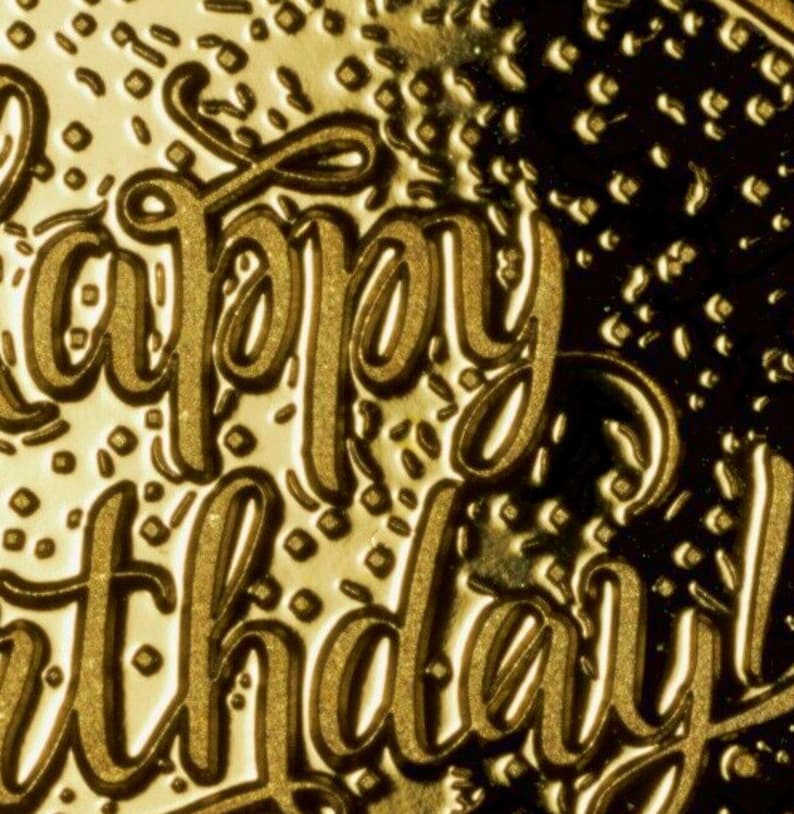 May include: A gold-coloured card with the words "Happy Birthday" embossed in a decorative script font. The background is textured with a pattern of small squares.