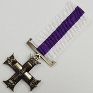 Pu&ograve; includere: Una medaglia a forma di croce d'argento con un nastro viola e bianco. La croce ha quattro bracci con una corona su ogni braccio. Il centro della croce presenta un motivo con una corona e una lettera 'R'.