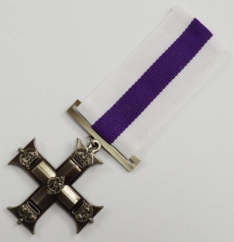 Pu&ograve; includere: Una medaglia a forma di croce d'argento con un nastro viola e bianco. La croce ha quattro bracci con una corona su ogni braccio. Il centro della croce presenta un motivo con una corona e una lettera 'R'.
