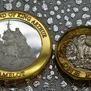 May include: A gold-colored coin with a silver center featuring a depiction of King Arthur on horseback with a sword. The coin has the text "The Legend of King Arthur" and "Camelot" around the edge. A standard British one pound coin is shown next to it.
