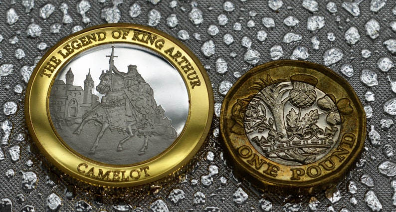 May include: A gold-colored coin with a silver center featuring a depiction of King Arthur on horseback with a sword. The coin has the text "The Legend of King Arthur" and "Camelot" around the edge. A standard British one pound coin is shown next to it.