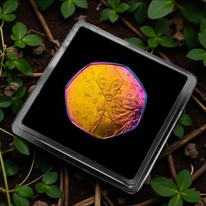 May include: A colorful coin featuring an alien figure and hieroglyphic-style symbols, displayed in a clear, square case. The coin has a rainbow iridescent finish, with hues of yellow, orange, pink, and purple. The case sits on a bed of soil and green plants.