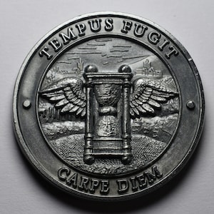 Op de afbeelding: Een zilverkleurige ronde munt met een zandloper, vleugels en de tekst "TEMPUS FUGIT CARPE DIEM".