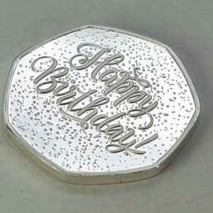 May include: A silver-colored coin with a raised inscription that reads "Happy Birthday!" The coin has a textured surface.