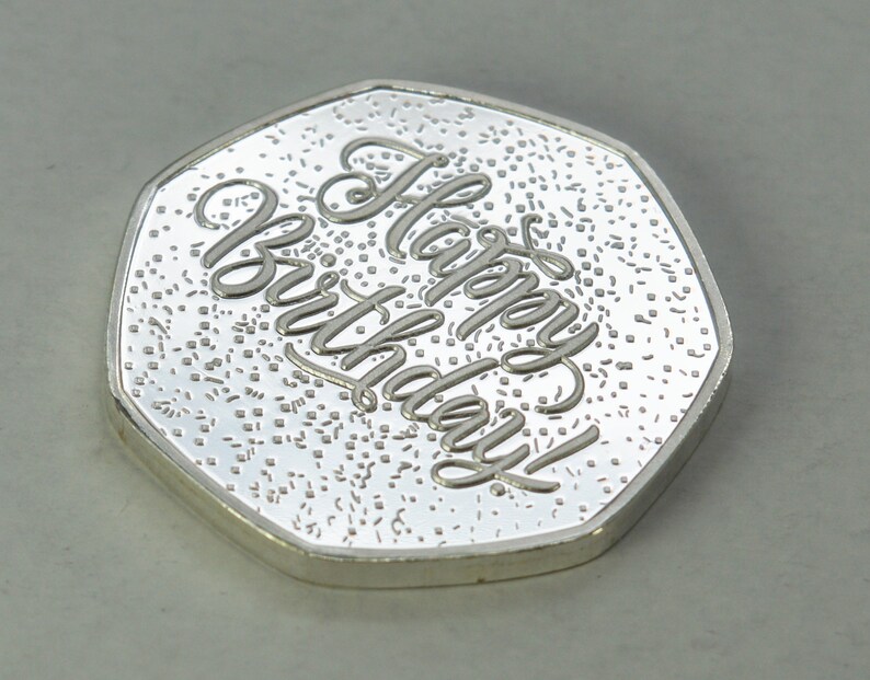 May include: A silver-colored coin with a raised inscription that reads "Happy Birthday!" The coin has a textured surface.