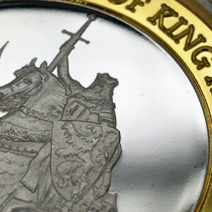 May include: A silver coin with a gold rim featuring a depiction of King Arthur on horseback, holding a sword. The inscription "King Arthur" is written in gold lettering around the rim.