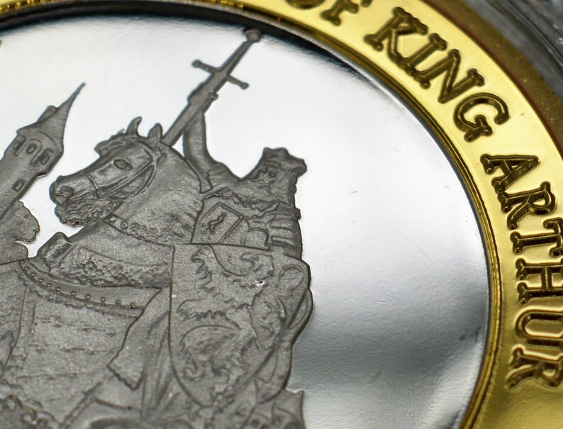 May include: A silver coin with a gold rim featuring a depiction of King Arthur on horseback, holding a sword. The inscription "King Arthur" is written in gold lettering around the rim.