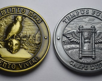 Op de afbeelding: Twee ronde, metalen munten met reliëfmotieven. De gouden munt heeft een uil die op een schedel zit met de tekst "Memento Mori" en "Memento Vivere". De zilveren munt heeft een gevleugelde zandloper met de tekst "Tempus Fugit" en "Carpe Diem".