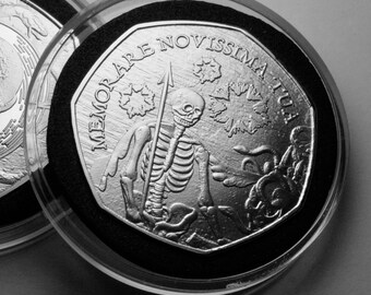 May include: A silver coin with a black and white image of a skeleton holding a spear. The coin has the text "MEMOARE NOVISSIMA TUI" around the edge.