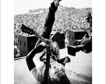 Puede incluir: Una fotografía en blanco y negro de un músico tocando la guitarra mientras está acostado de espaldas. El músico lleva una camisa blanca, pantalones negros y zapatos negros. El músico está actuando frente a una gran multitud.