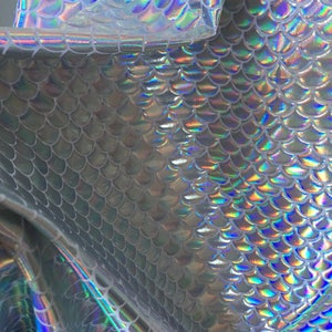 May include: Silver iridescent fabric with a mermaid scale pattern. The fabric is shiny and has a holographic effect.