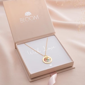 A gold family eternal ring necklace with three names engraved around the ring and three birthstones in the centre, in an open gift packaging box, with a foil gift card insert.