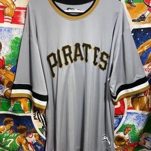 May include: A gray baseball jersey with the word "PIRATES" in black and gold lettering across the chest. The jersey has black and gold trim on the sleeves and collar. The jersey is hanging on a white hanger.