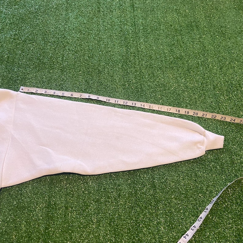 May include: A white long-sleeved garment sleeve is measured with a yellow measuring tape. The tape shows measurements up to 24 inches. The sleeve is on a green surface.