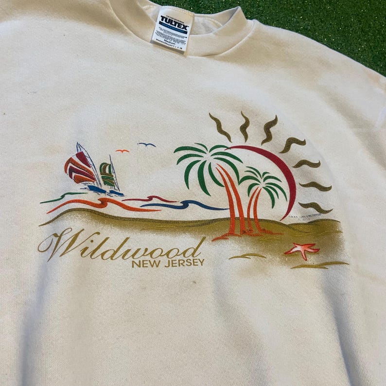 May include: Cream-colored sweatshirt featuring a beach scene with palm trees, sailboats, and the text "Wildwood NEW JERSEY". The graphic includes a red and brown sun, blue water, and a starfish.