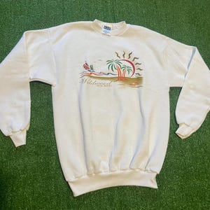 May include: White sweatshirt featuring a beach scene with palm trees, a sailboat, and a sun graphic. The text "Wildwood New Jersey" is printed below the graphic. The sweatshirt has long sleeves and a crew neck.