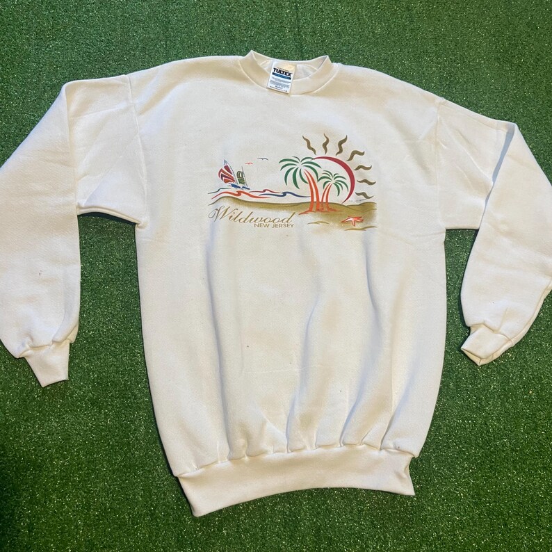 May include: White sweatshirt featuring a beach scene with palm trees, a sailboat, and a sun graphic. The text "Wildwood New Jersey" is printed below the graphic. The sweatshirt has long sleeves and a crew neck.