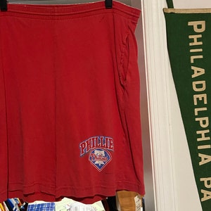 Può includere: Pantaloncini sportivi rossi con un logo blu e bianco dei Philadelphia Phillies. Un pennant verde con il testo "PHILADELPHIA PA" è appeso al muro dietro i pantaloncini.