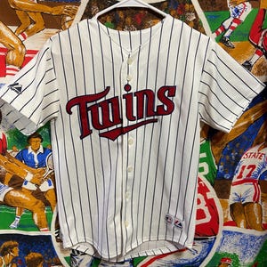 May include: A white baseball jersey with thin navy blue pinstripes, featuring the word Twins in red script across the chest. The button-down shirt has short sleeves, a Majestic logo on the right sleeve, and a team patch on the lower left front.