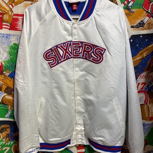 Op de afbeelding: Witte satijnen jas met het woord "SIXERS" in rode en blauwe letters over de borst. De jas heeft een blauw-rode gestreepte kraag, manchetten en tailleband. Voorzien van een knoopsluiting en twee voorzakken.