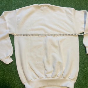 May include: A white crew neck sweatshirt with long sleeves. A measuring tape is laid across the chest of the sweatshirt, showing a measurement of approximately 21 inches. The sweatshirt is on a green surface.