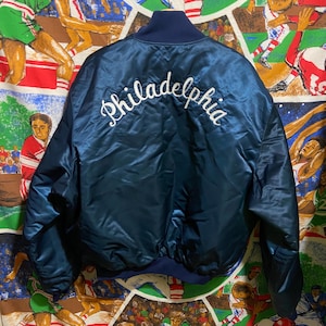 Peut inclure: Veste de bombardier en satin bleu marine avec le mot "Philadelphia" brodé en blanc sur le dos. La veste a un col et des poignets côtelés et une doublure matelassée. La veste est de style vintage.