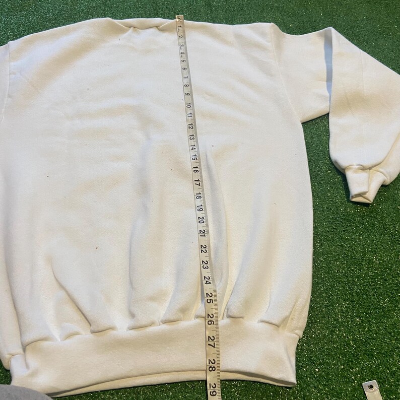 May include: A white sweatshirt with a crew neck and long sleeves. A measuring tape shows the length of the sweatshirt. The sweatshirt is on a green surface.