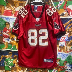 May include: A red football jersey with the number 82 in white on the front and shoulders. The jersey has black accents on the sleeves and collar, and the Buccaneers logo is visible on the sleeves. The jersey is hanging on a white hanger.