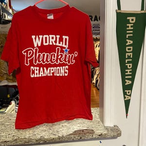 Puede incluir: Una camiseta roja de manga corta con el texto blanco WORLD PHUCKIN' CHAMPIONS impreso en la parte delantera. La palabra Phuckin' incluye una pequeña estrella azul sobre la letra i. La camiseta tiene cuello redondo y está en una percha rosa.