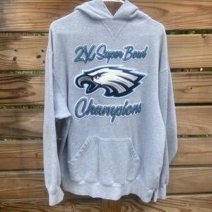 May include: A gray hooded sweatshirt with a front pocket and a drawstring hood. The sweatshirt features the text "2X Super Bowl Champions" in light blue script, along with a graphic of an eagle's head in white, black, and blue.