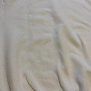 May include: Close-up of a cream-colored fabric with subtle variations in tone and texture. The fabric appears to be slightly wrinkled, with small dark specks scattered across the surface. The overall impression is of a soft, worn material.
