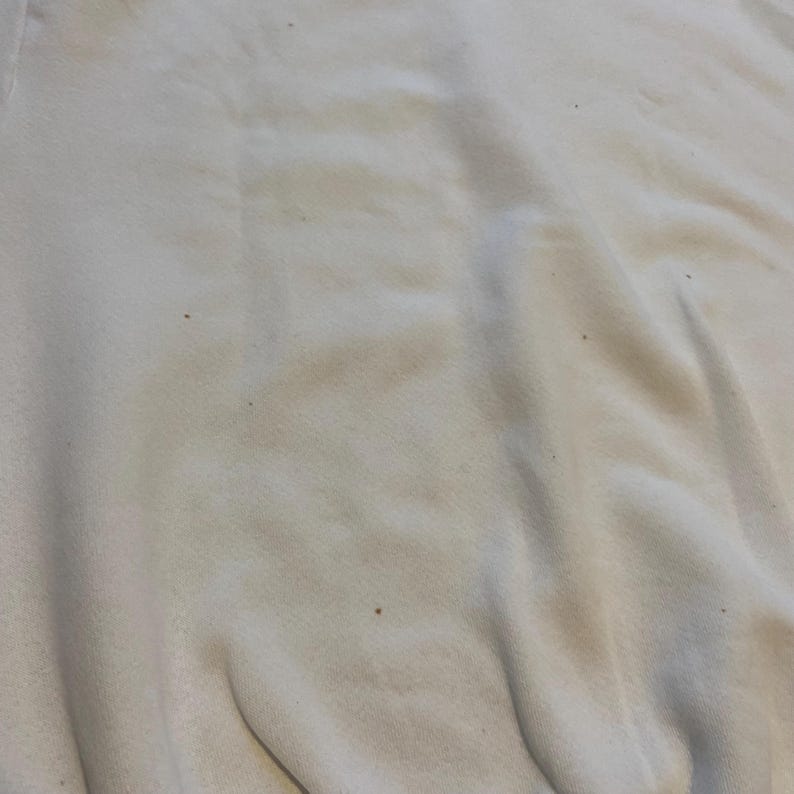 May include: Close-up of a cream-colored fabric with subtle variations in tone and texture. The fabric appears to be slightly wrinkled, with small dark specks scattered across the surface. The overall impression is of a soft, worn material.