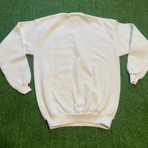 May include: A plain white sweatshirt with a crew neck and long sleeves. The cuffs and hem are ribbed. The garment is made of a soft, comfortable material, perfect for casual wear. The sweatshirt is displayed on a green surface.