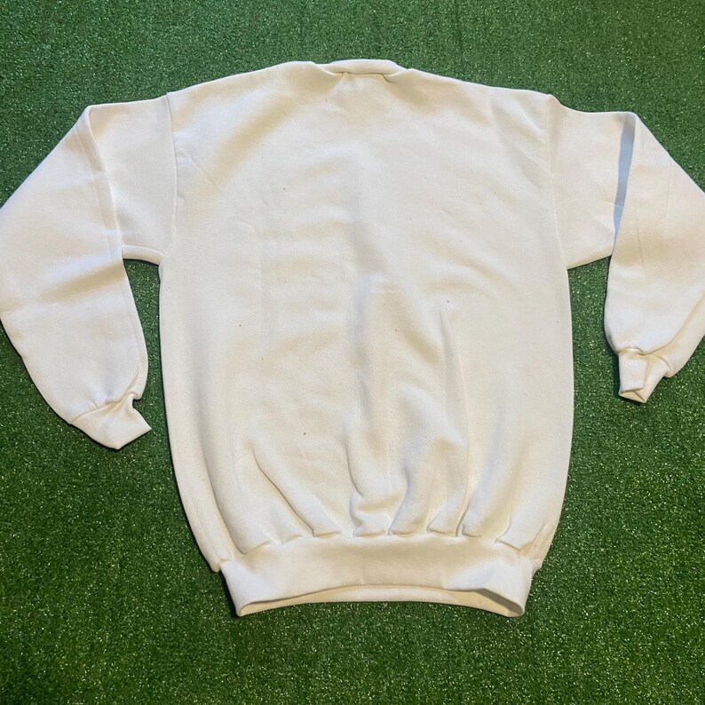 May include: A plain white sweatshirt with a crew neck and long sleeves. The cuffs and hem are ribbed. The garment is made of a soft, comfortable material, perfect for casual wear. The sweatshirt is displayed on a green surface.