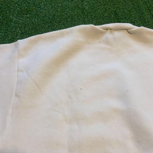 May include: Close-up of a cream-colored sweatshirt. The fabric appears soft and slightly worn, with subtle discoloration and small spots. The neckline and shoulder seam are visible, suggesting a casual style.
