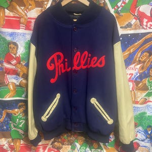 May include: A navy blue vintage Phillies baseball jacket with cream-colored sleeves. The word "Phillies" is embroidered in red script across the chest. The jacket has a button-up front and two front pockets with cream-colored trim.