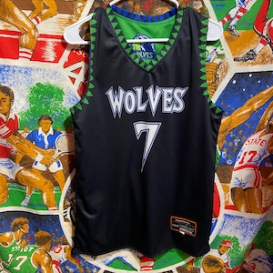 May include: Black basketball jersey with the word "WOLVES" in white script and the number "7" in white. The jersey has green and blue accents and a green triangle pattern along the edges. A white hanger holds the jersey.